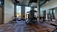 Located at 3000 Fast Ln in Fort Mill, SC, this contemporary fitness center is designed to support a full range of workout needs in a thoughtfully appointed, single-floor facility. The main gym area commands attention with dramatically soaring ceilings, exposed concrete columns, and floor-to-ceiling windows that flood the space with abundant natural light and outdoor views of the surrounding landscape. Patterned carpet tile flooring adds a modern aesthetic underfoot, while large wall-mounted mirrors visually expand the room and assist with form during strength training. The strength zone is comprehensively equipped with TKO multi-functional cable rigs, weight machines, a dumbbell rack, kettlebells, and a rowing machine. Cardio equipment, including multiple treadmills, is thoughtfully positioned along the window-lined wall, offering natural light and views while exercising. A separate studio space features smooth hardwood-style flooring, ceiling fans, linear LED lighting, full-length mirrors, wall-mounted TVs, and a barre bar—ideal for cycling classes, yoga, or group fitness sessions. Adjacent to the studio, a bright secondary exercise room is outfitted with spin bikes, stability balls, resistance tools, and direct outdoor access through glass-panel doors, creating a well-rounded and versatile fitness environment.