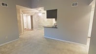 This single-floor space showcases an open-concept layout designed to maximize both functionality and flow. The main living area is anchored by notably high ceilings that lend a sense of spaciousness, complemented by soft, neutral-toned walls in a warm gray palette. Plush wall-to-wall carpeting covers the floors, adding comfort underfoot and contributing to the overall understated aesthetic.

A half-wall partition with a dark countertop breakfast bar separates the living area from the kitchen, offering a practical and visually appealing transition between the two spaces. The kitchen area features tile backsplash detailing visible behind the bar, adding a subtle textural element. A ceiling fan is centrally positioned in the living area, providing both air circulation and a functional overhead fixture.

A hallway leads toward additional rooms, with a white door visible at the far end, suggesting clear separation between living and private areas. HVAC venting is integrated into the upper wall, indicating a central climate control system. The overall ambiance is clean, neutral, and move-in ready, offering a versatile canvas adaptable to a variety of interior styles.