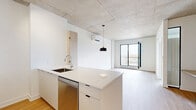 Located at 9450 Rue Meilleur in Montréal, this modern one-bedroom, one-bathroom unit offers a thoughtfully designed open-concept layout across a single floor. The combined kitchen and living area serves as the heart of the space, featuring light hardwood flooring, an exposed concrete ceiling, and a sleek kitchen island with a white quartz countertop and undermount sink. The kitchen is equipped with stainless steel appliances, including a full-sized refrigerator, stove, and built-in microwave, complemented by white cabinetry with light wood accents and a white tile backsplash. A wall-mounted air conditioning unit and large floor-to-ceiling black-framed windows and a glass balcony door in the living area allow for ample natural light and urban views of the Montréal cityscape. The bedroom is a bright, intimate space with light hardwood floors, large black-framed windows overlooking the city, and a baseboard heating unit. The bathroom is finished with large-format grey tiles on both the floor and walls, featuring a soaker tub with a glass shower enclosure and chrome fixtures, alongside a clean white vanity and toilet. A private balcony with glass railings extends the living space outdoors.