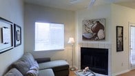 This single-story space showcases a welcoming living area designed with comfort and functionality in mind. The room centers around a classic tile-surround fireplace with a black firebox, serving as a natural focal point and adding warmth to the overall ambiance. Neutral-toned walls provide a clean, versatile backdrop that complements the space's understated aesthetic.

Natural light enters generously through a large window fitted with horizontal blinds, allowing for adjustable brightness throughout the day. A ceiling fan is installed overhead, contributing to year-round climate comfort. The textured ceiling adds a subtle architectural detail consistent with the room's practical design.

The living area accommodates a plush sectional sofa in a muted gray tone, paired with a glass-top coffee table that adds a touch of lightness to the arrangement. A classic floor lamp positioned near the window supplements the natural light with warm ambient illumination. Decorative wall art featuring earthy tones ties the room's color palette together cohesively. The flooring appears to be hardwood or wood-look material, adding warmth underfoot and enhancing the inviting character of the space.