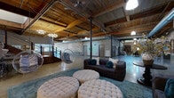 Located at 25 Draper St in Greenville, SC, this single-floor industrial loft space masterfully blends historic character with modern functionality. The expansive open-plan layout is defined by soaring ceilings clad in warm, rich wood planking supported by heavy timber beams and punctuated by bold structural steel columns — a hallmark of classic mill or warehouse architecture. Polished concrete floors run throughout, reflecting light and lending a sleek, contemporary contrast to the raw, textured surroundings.

Exposed brick walls, weathered and full of patina, line the perimeter and form dramatic arched openings that divide the space into distinct zones while maintaining an open, airy flow. Prominent exposed ductwork and pendant lighting reinforce the industrial aesthetic. A modern staircase with metal railings adds vertical interest and architectural flair.

The lounge area is warmly appointed with tufted ottomans, plush seating, and a hanging bubble chair, creating an inviting retreat within the larger space. A cozy media nook, enclosed by original brick walls and featuring a wall-mounted flat-screen TV, offers an intimate setting. Natural light filters through large windows near the reception area, brightening the otherwise moody interior. This versatile space is well-suited for events, creative studios, or a variety of commercial uses.
