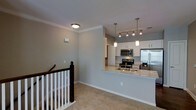This single-floor space showcases a thoughtfully designed open-concept layout that seamlessly connects the kitchen and living areas. The kitchen is a standout feature, equipped with stainless steel appliances including a French door refrigerator, electric range, and built-in microwave. White cabinetry provides ample storage, while a granite-topped peninsula with an undermount sink serves as both a functional workspace and a natural dividing element between the kitchen and the adjoining living space. Pendant lighting and a track light fixture above the peninsula add focused illumination and a contemporary aesthetic touch.

Crown molding runs along the ceiling throughout, lending a refined, polished character to the interior. The living area is finished with plush carpeting in neutral tones, contributing to a warm and comfortable atmosphere. Smooth, light-gray painted walls maintain a clean, modern look throughout the space.

A staircase with a classic dark wood banister and white spindles is prominently positioned, providing access to another level. The overall design balances practicality and style, creating an inviting environment suited to everyday living.
