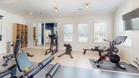 Located at 9001 Blue Flag Dr in Simpsonville, SC, this fitness center is a thoughtfully designed, single-floor facility offering a comprehensive workout experience. The space is divided into two visually distinct zones, each with its own character and purpose. The primary cardio area features bright white walls, light-toned flooring, and an abundance of natural light streaming through multiple windows fitted with white blinds. This section houses a row of treadmills, elliptical machines, and a rowing machine, along with a wall-mounted flat-screen television. Full-length mirrors are strategically placed to enhance the sense of openness throughout. The second zone features a striking navy blue shiplap accent wall and darker flooring, creating a more intense, performance-focused atmosphere. This area is equipped with a power rack loaded with weight plates, a full dumbbell rack, and an adjustable workout bench, catering to strength training needs. Track lighting with gold-toned fixtures illuminates both areas with a clean, modern aesthetic. Large mirrors in this zone further expand the visual space. The facility is accessible via a glass-panel door and includes clearly marked emergency exits, underscoring its community-use design. Together, the two zones provide a well-rounded, fully outfitted environment suitable for a wide range of fitness routines.