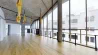 This expansive single-floor studio space showcases a compelling blend of industrial and contemporary design. Soaring ceilings with exposed concrete slab construction and visible overhead infrastructure create a raw, warehouse-inspired aesthetic throughout. The space is lined along one side by a continuous wall of floor-to-ceiling glass windows, flooding the interior with abundant natural light and offering open views of the surrounding urban streetscape.

Warm, light-toned hardwood flooring runs the full length of the studio, providing a striking visual contrast against the cool, grey concrete structural columns and ceiling. A sleek black metal railing system runs along the window-facing perimeter, adding a refined industrial detail to the space.

Suspended from the ceiling are multiple yellow aerial yoga hammocks or slings, indicating the space's use as a fitness or movement studio, specifically suited for aerial or suspension-based training. The generous open floor plan allows for flexible class formats and accommodates multiple participants comfortably.

The overall ambiance is bright, airy, and modern, with a distinctly urban industrial character that is both functional and visually engaging.