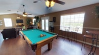 This single-floor recreational space is designed for leisure and entertainment, combining multiple activity zones into one cohesive layout. The centerpiece of the room is a full-sized pool table with a vibrant teal felt surface and solid wood frame, positioned prominently beneath a ceiling fan that helps maintain comfortable airflow throughout the space.

The room features warm-toned walls and hardwood-style flooring that lend a relaxed, inviting atmosphere. Natural light pours in through large windows along one wall and a set of glass-paned French doors, which also provide access to an outdoor area where additional bar-height seating and a wrought-iron table are visible.

Inside, a set of tall bar stools and a high-top table are positioned near the windows, creating a casual seating nook ideal for conversation or spectating. A popcorn machine adds a fun, social touch near the French doors. The lounge area in the background features comfortable seating alongside potted greenery, softening the space and adding a homey quality. Two ceiling fans ensure the room remains well-ventilated, making it a comfortable gathering spot for recreational activities.