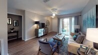 Located in Ridgeland, Mississippi, this single-story residence offers a well-designed layout that maximizes comfort and functionality throughout. The living room serves as the heart of the home, featuring wood-style flooring, a ceiling fan, and a sliding glass door that fills the space with natural light while providing direct access to an outdoor patio area. The open flow connects the living room to the dining area, which is defined by a pendant chandelier, a window with decorative curtains, and ample room for a full dining set. A breakfast bar with seating bridges the dining space to the galley-style kitchen, which is equipped with white cabinetry, granite-style countertops, a full suite of white appliances, and a double sink — all arranged efficiently along two parallel walls. The bedroom offers plush carpeting and direct access to a spacious walk-in closet with built-in shelving and hanging storage. The full bathroom features wood-style flooring, a white vanity with a granite-style countertop, a large mirror with overhead lighting, and a separate shower-tub area. Neutral gray tones are carried throughout the residence, creating a cohesive and calming ambiance from room to room.