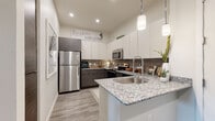 This thoughtfully designed single-floor residence showcases a cohesive modern aesthetic with clean lines and contemporary finishes throughout. The open-concept kitchen serves as a central highlight, featuring striking two-tone cabinetry — crisp white upper cabinets paired with dark wood-grain lower cabinets — complemented by granite countertops, a tile backsplash, and a full suite of stainless steel appliances including a refrigerator, range, microwave, and dishwasher. A peninsula with an integrated sink and pendant lighting above adds both functionality and visual appeal, creating a natural connection to the living area. The living space offers a welcoming atmosphere with wood-look flooring, generous wall space, and access to an outdoor area framed by dramatic floor-length drapery. Two well-proportioned bedrooms provide comfortable retreats, each featuring plush carpeting, large windows that invite natural light, and ample closet space. Both bathrooms are finished with modern floating-style vanities, white countertops, wood-grain cabinetry, and tub-shower combinations adorned with tasteful tile surrounds and wood-look flooring. The overall ambiance of the residence is fresh and contemporary, with a neutral color palette accented by warm and earthy tones throughout.