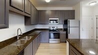 Located at 10 Newbridge Pkwy in Woodfin, North Carolina, this single-story unit offers a well-appointed layout with thoughtfully selected finishes throughout. The kitchen serves as a functional centerpiece, showcasing rich dark granite countertops paired with gray shaker-style cabinetry that extends to the ceiling for ample storage. Stainless steel appliances, including a range, microwave, and refrigerator, complement the light wood-look flooring that runs throughout the kitchen and connecting areas. A kitchen island provides additional counter space and a natural transition point into the living areas.

The living and bedroom spaces feature warm, neutral-toned carpeting and tall ceilings that lend an open, airy feel. A generously sized window in the bedroom allows natural light to filter in, offering views of the surrounding greenery. The neutral wall tones throughout create a clean, versatile backdrop.

The bathroom is finished with a gray vanity cabinet topped with a light stone-toned countertop, a undermount sink, and a modern multi-bulb light fixture above a large mirror. Light wood-look flooring continues here, tying the space together cohesively. A dedicated utility area includes wire shelving and washer/dryer hookups, adding everyday practicality to the overall design.