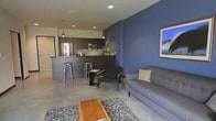 This single-floor space showcases a thoughtfully designed open-concept layout that seamlessly integrates the kitchen, dining, and living areas into one cohesive environment. The kitchen is outfitted with dark cabinetry, stainless steel appliances including a full-size refrigerator and built-in microwave, and a peninsula countertop with bar-height seating for two, making it both functional and social. Recessed and pendant lighting illuminate the kitchen with a modern, polished feel. The living area is anchored by a bold blue accent wall that adds depth and character to the space, complemented by neutral grey tones throughout. Polished concrete floors run continuously across the entire floor plan, contributing to the contemporary industrial aesthetic. The living room features a grey upholstered sofa, a sleek armchair, and a rustic wooden coffee table atop a textured area rug, creating a balanced mix of modern and warm design elements. Framed wall art adds a finishing touch. The open layout and cohesive color palette give the space a clean, urban feel.