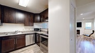 Located at 502 W Green St in Urbana, IL, this single-floor, 4-bedroom, 2-bathroom unit offers a practical and well-appointed layout. The kitchen is a standout feature, showcasing rich dark-wood cabinetry, granite-style countertops, and a full suite of stainless steel appliances including a dishwasher, electric range, and refrigerator, all set atop light wood-style flooring. The combined kitchen and living room area provides an open, functional gathering space anchored by a fireplace and accented with recessed lighting and light hardwood-style floors. A doorway from the living area provides convenient access to an in-unit stacked washer and dryer. The four bedrooms are modestly sized, with the carpeted rooms offering closets with wire shelving and windows fitted with horizontal blinds that allow natural light. Both bathrooms feature dark wood vanities with light countertops, large mirrors, tile flooring in an earthy stone pattern, and full tub-shower combinations. Two hallways connect the rooms efficiently throughout the single-floor layout. The overall aesthetic is clean and contemporary, with white walls and a consistent palette of dark wood and light neutral tones creating a cohesive, move-in-ready environment.