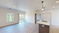 This well-designed single-floor residence showcases a clean, contemporary aesthetic with a thoughtful open-concept layout. The living area flows seamlessly from the kitchen, anchored by light-toned hardwood-style flooring that extends throughout the main spaces. Large sliding glass doors and adjacent windows flood the living room with natural light while providing views of the surrounding greenery, creating an airy and welcoming atmosphere.

The kitchen is appointed with rich, medium-toned wood cabinetry featuring sleek black hardware, complemented by marble-style stone countertops. A kitchen island with a built-in sink adds functional workspace, while pendant lighting overhead adds a stylish touch. Stainless steel appliances, including a French-door refrigerator, electric range, and over-the-range microwave, complete the well-equipped space.

The bedrooms are carpeted for comfort, each receiving natural light through sizable windows. The primary bedroom connects to a dedicated bathroom featuring a marble-style vanity countertop, wood cabinetry, a large mirror, and matte black accent fixtures. A spacious walk-in closet with wire shelving provides ample storage. A second bathroom mirrors the same refined finishes, maintaining a cohesive design throughout the residence.