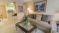 This single-floor residence exudes a warm and inviting ambiance, thoughtfully designed with classic elegance throughout. The living area serves as the heart of the space, centered around a plush, neutral-toned sofa adorned with decorative patterned pillows, complemented by a ornate glass-top coffee table with wrought iron detailing. Soft, ambient lighting from a tall floor lamp enhances the cozy atmosphere, while framed artwork on the walls adds a refined, curated character to the room. A lush floral arrangement on the coffee table introduces a touch of natural beauty, reinforcing the welcoming feel of the space. The warm, neutral color palette — carried across the walls, furnishings, and carpeted flooring — creates a cohesive and harmonious aesthetic throughout. Visible from the living area, the kitchen features bar-height seating, offering a convenient and casual dining option that connects the social spaces of the home. The open sightlines between the living and kitchen areas contribute to a sense of spaciousness, making this residence feel both functional and comfortable for everyday living.