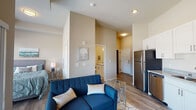 This single-floor unit showcases a thoughtfully designed open-concept layout with warm neutral tones and wood-look flooring throughout. The living area serves as the central hub of the space, featuring elevated ceilings that enhance the sense of openness. A sliding glass door floods the area with natural light and provides direct access to a private balcony with outdoor views, seamlessly connecting the interior to the exterior.

The kitchen runs along one wall in a galley-style configuration, outfitted with crisp white shaker-style cabinetry extending to the ceiling for generous storage. Granite-look countertops complement a full suite of stainless steel appliances, including a refrigerator, electric range, microwave, and dishwasher, along with a double-basin sink.

The sleeping area is integrated within the open layout, positioned near the balcony access and windows, allowing for natural light. A dedicated bathroom is accessible via a private corridor and features wood-look flooring, a white vanity with stone-look countertops, a walk-in shower with tiled surround, and modern chrome fixtures. The overall ambiance is clean, contemporary, and efficiently arranged for comfortable everyday living.