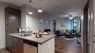 This 2-bedroom, 2-bathroom residence is thoughtfully designed with a modern aesthetic and efficient use of space throughout. The open-concept living and kitchen area serves as the heart of the home, featuring hardwood-style flooring, neutral gray walls, and a ceiling fan that adds to the comfortable ambiance. The kitchen is outfitted with dark espresso cabinetry, quartz-style countertops, stainless steel appliances including a refrigerator, range, and dishwasher, and a peninsula with pendant lighting that provides additional prep and dining space. The living area receives natural light through large windows and offers access to a private balcony, extending the living space outdoors. Both bedrooms are carpeted and feature walk-in closets with built-in shelving, providing ample storage. One bedroom includes a dedicated workspace, making it versatile for multiple uses. Each of the two bathrooms is finished with dark wood vanities, quartz-style countertops, large mirrors, and a tub-shower combination, with light wood-style flooring tying the design together. The overall aesthetic of this residence is contemporary and cohesive, balancing warm wood tones with cool gray accents for a polished, move-in-ready feel.