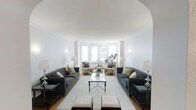 This single-floor space presents a sophisticated and well-appointed living room characterized by its graceful architectural design. A prominent arched entryway frames the room upon entry, immediately drawing attention to the space's classical sensibility. The ceiling is smooth and generously proportioned, contributing to an open, airy atmosphere throughout.

The living room is arranged with careful symmetry, featuring two charcoal-toned sofas positioned facing each other across a glass-topped coffee table, anchored by a plush, light-colored area rug. Complementing the seating arrangement are a pair of patterned accent chairs positioned near the bay window at the far end of the room, creating an inviting secondary seating area.

The bay window is a standout architectural feature, allowing abundant natural light to fill the interior and providing an open, welcoming ambiance. Warm hardwood flooring adds texture and richness to the overall aesthetic. Wall-mounted sconces and elegant table lamps provide layered lighting options. Crown molding along the upper walls reinforces the space's refined, classic character. The overall design is balanced, calm, and thoughtfully curated.
