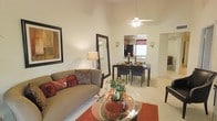 This single-story residence showcases an open-concept layout that connects the living and dining areas in a cohesive, flowing arrangement. The living area is highlighted by impressively high ceilings that amplify the sense of space, complemented by a ceiling fan with integrated lighting that adds both function and visual interest overhead. Neutral, warm-toned walls create a welcoming backdrop throughout, while light-colored tile flooring runs consistently across the space, enhancing brightness and continuity.

The living area comfortably accommodates a plush sofa adorned with decorative pillows, anchored by a circular glass coffee table atop a rich, terracotta-toned area rug. A large framed mirror and framed wall art add character and depth to the space. Wall sconces provide additional ambient lighting near the dining area.

The dining space features a dark-finished table surrounded by matching chairs, positioned beneath the ceiling fan and adjacent to a window that allows natural light to filter in. Beyond the dining area, a pass-through opening leads into the kitchen, which is visible in the background and benefits from additional natural light. The overall ambiance is warm, inviting, and thoughtfully designed for everyday living.