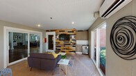This single-floor communal lounge space offers a well-appointed and thoughtfully designed interior that balances comfort with contemporary style. The layout is open and inviting, anchored by a distinctive multicolored wood-plank accent wall that serves as a striking focal point, housing a wall-mounted flat-screen television and a compact kitchenette with cabinetry, a mini refrigerator, and counter space — ideal for gatherings and casual entertaining.

The seating area features a charcoal-toned sofa accented with yellow throw pillows, complemented by modern side tables and a patterned area rug. A separate dining zone offers a glass-top table paired with contemporary lattice-style chairs, creating a functional space for group use. Additional modern seating is available throughout.

Floor-to-ceiling windows and multiple glass sliding and panel doors flood the interior with natural light while framing lush, landscaped outdoor views including mature palms, manicured hedges, and colorful plantings. A white beadboard ceiling enhances the bright, airy ambiance, while recessed lighting and a wall-mounted split air conditioning unit ensure year-round comfort. Tile flooring throughout adds durability and a clean aesthetic. Multiple access points provide seamless indoor-outdoor connectivity, making this space well-suited for community events and everyday social interaction.