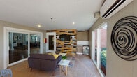 This single-floor communal lounge space offers a well-appointed and thoughtfully designed interior that balances comfort with contemporary style. The layout is open and inviting, anchored by a distinctive multicolored wood-plank accent wall that serves as a striking focal point, housing a wall-mounted flat-screen television and a compact kitchenette with cabinetry, a mini refrigerator, and counter space — ideal for gatherings and casual entertaining.

The seating area features a charcoal-toned sofa accented with yellow throw pillows, complemented by modern side tables and a patterned area rug. A separate dining zone offers a glass-top table paired with contemporary lattice-style chairs, creating a functional space for group use. Additional modern seating is available throughout.

Floor-to-ceiling windows and multiple glass sliding and panel doors flood the interior with natural light while framing lush, landscaped outdoor views including mature palms, manicured hedges, and colorful plantings. A white beadboard ceiling enhances the bright, airy ambiance, while recessed lighting and a wall-mounted split air conditioning unit ensure year-round comfort. Tile flooring throughout adds durability and a clean aesthetic. Multiple access points provide seamless indoor-outdoor connectivity, making this space well-suited for community events and everyday social interaction.