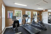 Located at 7843 Roswell Rd in Sandy Springs, GA, this fitness center is a well-equipped, single-floor exercise facility designed to support a comprehensive workout routine. The space features warm beige walls, a textured ceiling with bright fluorescent lighting, and durable dark rubber flooring throughout, creating a functional and motivating environment.

The cardio section is lined with several machines, including treadmills, ellipticals, and stationary bikes, positioned to take advantage of the natural light streaming through multiple windows. Those windows offer pleasant views of an outdoor pool area, providing an inviting backdrop during workouts. A large floor-to-ceiling mirror along one wall visually expands the space and allows users to monitor their form.

The strength and free weights area includes a multi-tier dumbbell rack stocked with a wide range of weights, as well as a stability ball for core training. Workout reference charts and fitness center rules — posted in both English and Spanish — are displayed along the walls for easy reference.

The space is accessed through a centrally located door flanked by windows, contributing to its bright, open feel. Safety features include clearly marked emergency exits, a fire extinguisher, and a security camera. This fitness center provides a complete and accessible workout facility for those in the Sandy Springs community.