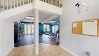 Located in Stuart, Florida, this fitness center offers a comprehensive exercise facility designed to support a wide range of workout routines. The space is finished with bold teal accent walls and durable rubber flooring, creating an energetic and functional environment. Skylights and flush-mounted ceiling fixtures provide ample illumination throughout the open floor plan.

The strength training area is well-appointed, featuring a multi-station cable machine, a Smith machine with an adjustable bench, and a full dumbbell rack organized along a floor-to-ceiling mirrored wall, allowing users to monitor their form with ease. A dedicated TRX Training Zone lines one wall, complete with suspension trainers, kettlebells, medicine balls, and a plyometric box, catering to functional and high-intensity training.

The cardio section includes a treadmill, elliptical, stationary bike, and air bike, all positioned near windows that offer direct views of the outdoor pool and surrounding palm trees — bringing a refreshing Florida ambiance to every workout. A stability ball and additional free-weight accessories round out the available equipment.

The facility also connects to an adjacent tiled entryway with a staircase, suggesting shared amenity access. Ventilation is provided through ceiling-mounted HVAC vents, ensuring comfort throughout the year in the warm South Florida climate.