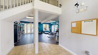 Located in Stuart, Florida, this fitness center offers a comprehensive exercise facility designed to support a wide range of workout routines. The space is finished with bold teal accent walls and durable rubber flooring, creating an energetic and functional environment. Skylights and flush-mounted ceiling fixtures provide ample illumination throughout the open floor plan.

The strength training area is well-appointed, featuring a multi-station cable machine, a Smith machine with an adjustable bench, and a full dumbbell rack organized along a floor-to-ceiling mirrored wall, allowing users to monitor their form with ease. A dedicated TRX Training Zone lines one wall, complete with suspension trainers, kettlebells, medicine balls, and a plyometric box, catering to functional and high-intensity training.

The cardio section includes a treadmill, elliptical, stationary bike, and air bike, all positioned near windows that offer direct views of the outdoor pool and surrounding palm trees — bringing a refreshing Florida ambiance to every workout. A stability ball and additional free-weight accessories round out the available equipment.

The facility also connects to an adjacent tiled entryway with a staircase, suggesting shared amenity access. Ventilation is provided through ceiling-mounted HVAC vents, ensuring comfort throughout the year in the warm South Florida climate.