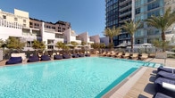 Located at 825 S Hill St in the heart of Downtown Los Angeles, this outdoor amenity deck offers a resort-inspired escape amid the urban skyline. The space centers around a generously sized swimming pool with crystal-clear blue water, surrounded by a spacious sun deck finished in smooth, neutral-toned concrete pavers. An array of lounge chairs and cushioned seating — including plush bean-style loungers shaded by large umbrellas — provide ample space for relaxation under the Southern California sun. Tall palm trees punctuate the deck, adding a lush, tropical character to the setting. A dedicated lap pool runs along one edge of the terrace, enclosed by sleek glass railings that open up to sweeping views of the Downtown LA skyline, including iconic high-rises and historic commercial buildings. A standout architectural feature is a boldly patterned, ornate tile wall that anchors one corner of the deck, introducing an artistic, Mediterranean-influenced accent to the contemporary design. The deck is framed by a striking modern tower above, reinforcing the space's urban sophistication. Overall, the amenity deck blends thoughtful design, functional amenities, and dramatic city views into a cohesive and inviting outdoor retreat.
