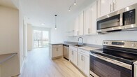 This single-floor unit presents a clean, contemporary design characterized by light hardwood-style flooring, crisp white walls, and an open, airy layout. The kitchen is a standout feature, outfitted with white shaker-style cabinetry, grey countertops, and a full suite of stainless steel appliances including an electric range, over-the-range microwave, dishwasher, and refrigerator. A single-basin sink, open shelving, and track lighting add both functionality and style, while a pendant light signals the transition toward the living area. The living space connects seamlessly to a private balcony accessed through full-height sliding glass doors, which flood the interior with natural light and offer open views of the surrounding urban skyline. The bedroom is comfortably sized with light-toned flooring and direct access to both the kitchen area and the bathroom. The bathroom maintains the unit's consistent aesthetic with white cabinetry, a white countertop vanity, a large mirror, and a shower-tub combination. Throughout the space, the neutral palette, modern hardware, and abundant natural light create a cohesive, understated urban living environment.