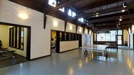This distinctive single-floor space masterfully combines industrial character with recreational functionality. The architecture is immediately striking, defined by bold exposed dark timber beams and trusses that span the high ceiling, lending a rustic yet refined aesthetic. Clerestory windows line the upper walls, drawing in abundant natural light and creating an airy, open atmosphere despite the space's substantial footprint.

The main open area features a smooth, polished concrete floor in a cool grey tone, providing a durable and versatile surface well-suited for a variety of activities. A ping-pong table anchors the leisure zone, underscoring the space's focus on recreation and social engagement.

Flanking the main floor is a dedicated fitness area, partially enclosed by a low plywood partition with dark framing. Through the openings, cardio equipment including a treadmill is visible, offering a functional workout zone integrated seamlessly into the overall layout.

The walls feature textured white paneling with an organic, sculpted quality, adding a bold artistic element that contrasts with the raw structural materials overhead. Warm wall-mounted light fixtures complement the natural light, creating an inviting and energetic ambiance throughout. This space is ideally suited for community recreation, fitness, and social activities.