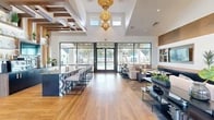 Located in Jacksonville, FL, this thoughtfully designed community clubhouse delivers a sophisticated and welcoming atmosphere across a generously sized single-floor layout. The main gathering area is anchored by soaring ceilings with dramatic raised clerestory windows that flood the interior with abundant natural light. A striking gold tiered chandelier serves as the centerpiece overhead, complementing the warm-toned hardwood flooring that flows throughout the space.

A sleek bar-height counter with a bold geometric-patterned base and plentiful seating offers an inviting social hub, while floor-to-ceiling black-framed glass doors open directly to views of an outdoor pool and surrounding community. The lounge area features a built-in wood-paneled media wall with integrated shelving and dual flat-screen displays, paired with a plush sectional sofa and a geometric-patterned area rug that adds texture and warmth.

A dedicated co-working nook provides a quieter retreat, outfitted with private booth-style blue upholstered seating, individual tables, and modern pendant lighting — ideal for focused work or casual meetings. Additional lounge seating zones with rounded accent chairs and wall-mounted sconce lighting create versatile spaces for both socializing and relaxation. The overall design blends contemporary finishes with warm natural wood accents, resulting in a polished and functional communal environment.