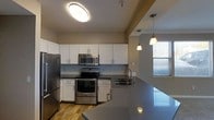 This single-story unit is designed with a functional and modern layout. The kitchen serves as a central highlight, featuring stainless steel appliances including a refrigerator, electric range, and over-the-range microwave. White shaker-style cabinetry extends to the upper walls, providing generous storage, while dark countertops offer a contrasting visual element. A large kitchen island with a built-in sink doubles as a breakfast bar, illuminated by stylish pendant lighting. Wood-look flooring runs throughout the kitchen, adding warmth to the space. A window allows natural light to filter in, brightening the area.

The living area is finished with neutral-toned walls and wall-to-wall carpeting, creating a comfortable and inviting atmosphere. A ceiling fan is installed overhead, contributing to air circulation and comfort. Crown molding details and a tray ceiling add architectural interest to the room. A corridor connects the living space to additional areas of the unit, with a window at the far end drawing in natural light. HVAC vents are integrated into the ceiling, ensuring efficient climate control throughout.