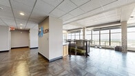 Located on N Waco St in downtown Wichita, Kansas, the 24th Floor is a spacious, single-level commercial space offering a generously sized open-plan layout. The space is defined by floor-to-ceiling windows that wrap around the perimeter, flooding the interior with natural light and providing sweeping panoramic views of the Wichita skyline and surrounding cityscape.

The flooring throughout features large-format stone-look tile in a cool metallic tone, complemented by areas of darker, warm-toned tile that help define distinct zones within the open layout. Exposed cylindrical ductwork runs along the upper portion of the space, lending an industrial character that contrasts with the clean drop-tile ceilings found in other sections. Structural brick and concrete columns punctuate the floor plan, adding architectural texture.

Flexible seating arrangements include high-top bar tables with modern chairs and green upholstered booth seating, supporting both collaborative and individual work. A curved wood-paneled reception or service station serves as a central anchor point, accompanied by a wall-mounted display screen. A more open, multipurpose area at one end provides additional event or gathering capacity. Elevator access is conveniently integrated into the space. The overall ambiance is contemporary and professional, with an energetic, collaborative character well-suited for co-working, events, or professional gatherings.