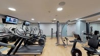 The Fitness Studio at 1 St. Mary's Convent in Peekskill, NY is a thoughtfully designed, single-floor exercise facility offering a comprehensive range of workout equipment for residents and users alike. The studio features light hardwood-style flooring throughout, neutral textured walls with a classic gray accent border, and recessed lighting complemented by ceiling-mounted HVAC units that ensure a comfortable environment year-round.

The cardio zone is well-appointed with a row of treadmills, elliptical machines, stationary bikes, and stair-climbers, several of which are equipped with individual display screens for interactive workouts. A wall-mounted flat-screen TV is positioned for easy viewing during cardio sessions. The strength and resistance training area includes a multi-station cable machine, a dedicated weight bench with an adjustable incline, and a full rack of dumbbells ranging across multiple weight increments. A medicine ball rack along the wall adds versatility for functional training routines.

Large mirrors are strategically placed to expand the sense of space and allow users to monitor their form. The studio is accessible through a frosted glass door and maintains a clean, organized layout that maximizes usable floor space. A wall clock and climate controls add to the practicality of the space, making the Fitness Studio a fully functional and inviting environment for a wide range of workout needs.