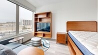 Located at 2132 Stokes Blvd in Cleveland's University Circle neighborhood, this efficiently designed one-bedroom, one-bathroom unit makes excellent use of its compact footprint. Light wood-look flooring runs throughout, lending a clean, cohesive aesthetic to the entire space.

The open living area connects seamlessly to a modern kitchen outfitted with gray cabinetry, a stainless steel refrigerator, built-in microwave, and white countertops. A large window at the living end of the unit fills the space with natural light while offering views of the surrounding Cleveland cityscape.

The bedroom is furnished with warm walnut-toned pieces, including a platform bed, a matching nightstand, and a built-in shelving unit with an integrated media center. A colorful area rug and a stacked pouf add a touch of personality to the space.

A stacked washer and dryer unit is conveniently tucked behind a closet door, providing in-unit laundry without sacrificing valuable living space. The bathroom features a clean, contemporary design with a gray vanity, white countertop, large mirror, a tub-shower combination, and matte black hardware and grab bar accents throughout.

Overall, the unit presents a modern, functional design well-suited for urban living in Cleveland, OH.