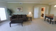 This single-floor space offers a welcoming and functional layout, highlighted by soft sage-green walls and neutral beige carpeting that create a calm, cohesive atmosphere throughout. The living area is generously proportioned, providing ample room for comfortable seating and furnishings. Crown molding runs along the ceiling perimeter, adding a refined architectural detail that elevates the overall aesthetic. A ceiling vent and smoke detector are discretely integrated into the ceiling, reflecting practical design considerations.

An elegant arched doorway serves as a transitional element, connecting the living area to adjacent spaces and lending a sense of architectural character to the interior. Through this archway, a six-panel door is visible, alongside a partial view of a secondary room featuring dark wood cabinetry and what appears to be a bathroom vanity area, suggesting well-appointed fixtures and storage.

The living space is bathed in ambient light, enhanced by a table lamp that contributes a warm glow to the room. Framed wall art adds a decorative touch, while the overall design strikes a balance between comfort and understated elegance, making this a practical yet inviting living environment.