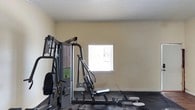 Located at 190 N Corry Field in Pensacola, Florida, this single-floor fitness facility provides a functional and focused workout environment. The exercise space features neutral-toned walls that create a clean, distraction-free atmosphere conducive to training. Dark rubber flooring covers the workout area, offering durability and safety underfoot — a practical choice for a high-use fitness setting. The room is equipped with a multi-station strength training machine, allowing users to perform a variety of resistance exercises in one location. A window fitted with horizontal blinds allows natural light to filter into the space, brightening the interior while maintaining privacy. The textured ceiling adds a classic architectural finish to the room. The layout is straightforward and efficient, maximizing the usable floor space for equipment and movement. Overall, this facility presents a no-frills, purpose-built environment designed to meet the essential fitness needs of its users, making it a practical resource for those seeking a convenient and accessible place to exercise in the Pensacola area.