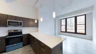 This single-floor space showcases a contemporary open-plan layout that seamlessly integrates the kitchen and living area into one cohesive environment. The kitchen is outfitted with dark espresso-toned cabinetry, providing a striking contrast against the bright white countertops that extend across both the perimeter workspace and a generously sized central island. The island serves as a natural focal point, offering additional storage and functioning as an ideal space for casual dining or entertaining. Stainless steel appliances, including a built-in microwave and range, add a polished, functional element to the kitchen design.

Soaring ceilings amplify the sense of openness throughout, while exposed industrial elements — including track lighting and overhead piping — lend a loft-like character to the space. Pendant lighting hangs above the island, providing both task illumination and aesthetic warmth. The polished concrete flooring ties the design together with an urban, modern aesthetic. A large, multi-pane window in the living area floods the space with natural light and frames views of the surrounding cityscape, enhancing the overall ambiance of this thoughtfully designed urban space.