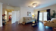 This single-floor residence offers a cohesive and functional layout finished with rich, dark hardwood-style flooring that runs throughout the entire space, creating a warm and unified aesthetic. The living room serves as the heart of the home, featuring elegant crown molding, a ceiling fan with light fixture, and a set of French doors that invite natural light and provide outdoor access. The room comfortably accommodates a seating area and opens to the rest of the unit with ease.

The galley-style kitchen is lined on both sides with deep, rich cherry wood cabinetry and granite-look countertops, offering generous storage and prep space. A full-size refrigerator and a sink with ample counter space make this kitchen both practical and visually appealing.

The primary bedroom is a peaceful retreat, with large draped windows that filter soft natural light, while a second bedroom provides flexible living space, currently configured with bunk beds and dark wood furniture, making it well-suited for multiple occupants or guests.

The bathroom features a wide vanity with white cabinetry, a large mirror, and hardwood-style flooring that continues the home's consistent design language. A dedicated in-unit laundry area with a full-size washer and utility shelving adds everyday convenience to this thoughtfully designed residence.
