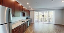 This well-appointed single-floor unit offers a thoughtfully designed open-concept layout that seamlessly connects the kitchen and living areas. The kitchen is a standout feature, showcasing rich dark cherry wood cabinetry extending to the ceiling, light granite countertops, a stainless steel appliance suite including a refrigerator, electric range, microwave, and dishwasher, and a undermount sink — all set atop warm hardwood-style flooring that runs throughout the entire unit. The spacious living area is bathed in natural light, thanks to large sliding glass doors that open onto a private balcony overlooking mature trees, creating a serene outdoor connection. An additional window further enhances the bright and airy ambiance of the space. The unit includes multiple bedrooms, each featuring hardwood-style flooring, windows for natural light, and closet storage. The bathroom is cleanly finished with a dark wood vanity, white countertop, a large mirror, tile flooring, and a full soaking tub with subway tile surround. Recessed lighting throughout adds a modern touch, while neutral gray walls provide a versatile backdrop. The overall aesthetic is clean, contemporary, and move-in ready.