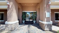 This single-story space makes a memorable first impression with a stately exterior entrance framed by a covered portico, stone-accented walls, and large glass double doors flanked by tall potted plants. A paved walkway leads visitors toward the building, with palm trees visible in the background, hinting at a warm, sunny locale.

Inside, the grand lobby immediately commands attention with its dramatic rotunda design, featuring soaring ceilings that rise to a clerestory of windows, flooding the space with natural light. A coffered ceiling detail crowns the circular focal point, from which a classic chandelier is suspended. At the center of the lobby stands an ornate round glass-topped table adorned with a lush floral arrangement, reinforcing the space's refined character.

Large, mature indoor trees in stone planters anchor each side of the lobby, lending a warm, organic balance to the architecture. Light-toned tile flooring with an inlaid decorative medallion adds an elegant finishing touch underfoot. Neutral, warm-toned walls and arched architectural details frame the view toward the interior corridor, where additional natural light pours in through the far entryway. The overall ambiance is welcoming, polished, and professionally appointed, suggesting a high-end commercial or hospitality use.
