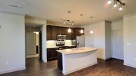 Located in North Richland Hills, Texas, this single-floor residence offers a thoughtfully designed open-concept layout that seamlessly blends functionality with modern aesthetics. The kitchen serves as the centerpiece of the space, showcasing rich dark wood cabinetry that extends to the upper walls, providing ample storage while creating a striking visual contrast. Light granite countertops run throughout, including a generously sized curved island with an integrated sink — ideal for meal prep and casual dining. Stainless steel appliances, including a gas range, built-in microwave, and French door refrigerator, complete the chef-ready setup. Pendant lighting hangs elegantly above the island, complemented by a track lighting system that illuminates the cooking area. The kitchen opens fluidly into the living area, creating an expansive, connected feel. Rich dark hardwood flooring flows throughout, anchoring the space with warmth and continuity. A hallway visible from the main living area leads to additional rooms, hinting at a well-organized floor plan. The neutral wall tones and clean white trim provide a versatile backdrop that suits a variety of interior styles. Overall, the space delivers a polished, contemporary living environment with quality finishes throughout.