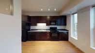 This single-floor unit presents a clean, modern design with a practical layout that maximizes functionality across its living spaces. The kitchen serves as a standout feature, showcasing rich dark espresso cabinetry extending to the ceiling, complemented by sleek dark countertops and a full suite of black appliances, including a refrigerator, electric range, built-in microwave, and dishwasher. A double basin sink is positioned beneath under-cabinet lighting, while track lighting overhead provides focused illumination. Light hardwood-style flooring runs through the kitchen and main living areas, adding warmth to the space.

The bedroom is carpeted for comfort and features a distinctive interior window that connects visually to the adjacent living area, allowing borrowed light to filter through. A wall-mounted HVAC vent and recessed lighting provide climate control and illumination. A private door leads from the bedroom to additional spaces.

The bathroom is finished in a crisp, contemporary style with a dark vanity topped by a white countertop and an undermount sink with chrome fixtures. A large frameless mirror is illuminated by a multi-bulb vanity light bar, creating a bright, functional grooming area. Neutral wall tones throughout the unit contribute to a cohesive and understated aesthetic.