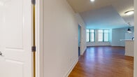 This well-appointed single-floor residence offers a clean, modern aesthetic throughout, characterized by warm hardwood flooring, crisp white walls, and an abundance of natural light. The open-concept living area is anchored by floor-to-ceiling windows fitted with horizontal blinds, framing views of the surrounding cityscape and filling the space with daylight. A wide hallway connects the entryway to the main living space, providing a smooth and intuitive flow through the unit. The kitchen is thoughtfully designed with white shaker-style cabinetry extending to the ceiling, stainless steel appliances including a side-by-side refrigerator with ice maker, a built-in microwave, and a range. A peninsula with a white countertop and pendant lighting overhead creates a natural boundary between the kitchen and the dining and living areas. The bedroom is generously proportioned, featuring its own large window with city views and direct access to a closet. The en-suite bathroom is finished with warm beige tile flooring and tub surround, white cabinetry, and a granite-topped vanity. A second full bathroom mirrors this finish with similar tile work, granite counters, and clean white fixtures. Recessed lighting throughout ensures consistent illumination across all areas of the residence.