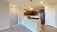 Located at 9295 One Deerfield Pl in Mason, Ohio, this single-floor residence offers a thoughtfully designed layout with contemporary finishes throughout. The kitchen serves as a central highlight, featuring rich dark-toned cabinetry, a full suite of stainless steel appliances including a refrigerator, range, and microwave, and a light-colored countertop peninsula with an integrated sink. Stylish globe pendant lights add a modern touch, while wood-look flooring grounds the space with warmth and durability. The open configuration allows the kitchen to connect seamlessly to the adjacent living areas, which are finished with plush carpeting and neutral wall tones. Bedrooms are generously proportioned, with large windows and sliding glass doors that invite natural light and provide outdoor views. Ceiling fans provide year-round comfort. The primary suite connects to a well-equipped en-suite bathroom featuring a dual-sink vanity with dark cabinetry, a glass-enclosed walk-in shower, and wood-look flooring. An additional bathroom offers a soaking tub with tile surround. A spacious walk-in closet with multi-tiered wire shelving provides ample storage. The overall ambiance is clean, modern, and move-in ready.