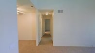 This single-floor residence is designed with a straightforward and functional layout, centered around a carpeted hallway that connects the various rooms. The interior features neutral white walls throughout, creating a clean and cohesive aesthetic that allows for easy personalization. Wall-to-wall carpeting in a warm beige tone runs through the main living area and hallway, adding a sense of comfort underfoot. The hallway is lined with doorways leading to additional rooms, and a ceiling-mounted flush light fixture provides consistent illumination along the corridor. HVAC vents are positioned both on the ceiling and upper walls, ensuring efficient climate control throughout the space. The open transition between the main living area and the hallway enhances the sense of flow and connectivity within the home. The high ceilings contribute to an airy, open atmosphere, making the interior feel spacious despite the compact footprint. Overall, the space presents a move-in-ready environment with a simple, understated design that offers a versatile canvas for a variety of interior styles.