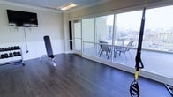 This single-floor fitness space combines functionality with an open, airy atmosphere. The room is characterized by rich, dark hardwood flooring that extends throughout, lending a polished and durable surface well-suited for exercise activity. Crown molding and wainscoting along the lower walls add a refined architectural touch, elevating the space beyond a typical gym environment.

Dominating one wall is an expansive bank of large sliding glass windows and a glass door that floods the interior with abundant natural light. These floor-to-ceiling windows offer sweeping views of the surrounding urban landscape, creating a bright and motivating workout environment. The glass door provides direct access to an outdoor terrace, which is furnished with a table and chairs — ideal for rest and recovery in the open air.

Overhead fluorescent lighting ensures the space remains well-lit regardless of the time of day. Fitness equipment, including an adjustable weight bench, is positioned to take advantage of the open floor plan, allowing for flexible arrangement of workout stations. The combination of natural light, outdoor access, city views, and tasteful interior finishes makes this a distinctive and practical fitness facility.