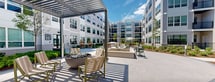 Located at 6 Becker Farm Rd in Roseland, NJ, this thoughtfully designed outdoor amenity courtyard is set within a modern multi-story residential community. The space features a large open-air patio with smooth, light-toned pavers that serve as the central gathering area, surrounded by a variety of seating arrangements including bistro tables, Adirondack-style chairs, and lounge seating to accommodate both social gatherings and quiet relaxation. A standout feature is the circular fire pit area, furnished with green Adirondack chairs arranged to encourage conversation and community. A covered pergola provides shaded seating, while an outdoor grilling station with bar-height tables offers a dedicated space for outdoor dining. Manicured landscaping throughout the courtyard includes mulched planting beds filled with ornamental shrubs, flowering plants, and young shade trees, contributing to a vibrant and green atmosphere. A section of lush artificial turf adds a recreational lawn element, ideal for casual outdoor activities. Bollard lighting is integrated throughout the pathways, ensuring the space is functional after dark. The courtyard is enclosed by the surrounding residential building, featuring contemporary gray siding, large windows, and private balconies, creating a cohesive and private community retreat under open skies.