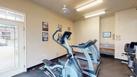 This fitness center is a compact, single-room facility designed to provide a well-rounded workout experience. The room features dark rubber flooring throughout, offering both durability and safety during exercise. Cardio equipment is a highlight of the space, with multiple treadmills and elliptical machines arranged to maximize the available floor area. A dedicated free weights section lines one wall, offering a selection of kettlebells and dumbbells alongside resistance training accessories. A full-width wall mirror on one side visually expands the room while allowing users to monitor their form during workouts. Natural light filters in through a large window with horizontal blinds, complementing the overhead fluorescent lighting and a ceiling fan that helps maintain air circulation. A wall-mounted flat-screen television provides entertainment during workouts. The room also includes a built-in utility sink with a granite countertop in an alcove, adding a practical touch for hygiene. Motivational framed prints and fitness center rules are displayed on the walls. The overall ambiance is functional and straightforward, offering an efficient and accessible fitness environment.