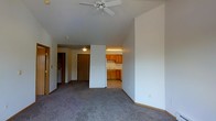 This single-floor unit features an open and airy layout connecting the main living area to the kitchen. The living space is finished with wall-to-wall carpet and benefits from vaulted ceilings that enhance the sense of height and openness. A ceiling fan is centrally mounted, providing both lighting and air circulation throughout the room. Neutral-toned walls keep the space bright and versatile.

The kitchen is partially open to the living area, separated by a partial wall that maintains a sense of connection between spaces. It is equipped with warm-toned wooden cabinetry, a white refrigerator, and under-cabinet lighting that illuminates the countertops and workspace. A sink is visible along the kitchen wall, and the kitchen flooring transitions from the carpeted living area to a hard surface, clearly delineating the two zones.

A hallway extends from the main living area toward what appears to be additional rooms, with a wood-toned door visible at its end. A separate door, also visible from the living area, likely serves as the unit's entry point. The overall ambiance is functional and straightforward, with a clean, modest design suitable for everyday living.