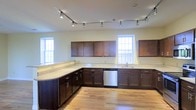 This single-floor space showcases a generously sized kitchen as its centerpiece, designed with both functionality and aesthetics in mind. Rich, dark wood cabinetry lines the walls in an L-shaped configuration, providing abundant storage throughout. The upper and lower cabinets are complemented by light-toned granite countertops that offer extensive prep and workspace, including a curved peninsula that adds additional surface area and visual interest.

A stainless steel dishwasher is seamlessly integrated into the cabinetry, contributing to the kitchen's modern, cohesive look. The sink is positioned beneath one of several double-hung windows, which flood the space with natural light and lend an open, airy feel to the interior. The light hardwood flooring runs throughout, adding warmth and continuity to the space.

Overhead, a track lighting system runs along the ceiling, providing adjustable, directed illumination across the entire kitchen area. The neutral wall tones work harmoniously with the dark cabinetry and light countertops, creating a balanced and inviting atmosphere. Overall, the space presents a well-appointed, practical layout suited for everyday use.