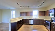 This single-floor space showcases a generously sized kitchen as its centerpiece, designed with both functionality and aesthetics in mind. Rich, dark wood cabinetry lines the walls in an L-shaped configuration, providing abundant storage throughout. The upper and lower cabinets are complemented by light-toned granite countertops that offer extensive prep and workspace, including a curved peninsula that adds additional surface area and visual interest.

A stainless steel dishwasher is seamlessly integrated into the cabinetry, contributing to the kitchen's modern, cohesive look. The sink is positioned beneath one of several double-hung windows, which flood the space with natural light and lend an open, airy feel to the interior. The light hardwood flooring runs throughout, adding warmth and continuity to the space.

Overhead, a track lighting system runs along the ceiling, providing adjustable, directed illumination across the entire kitchen area. The neutral wall tones work harmoniously with the dark cabinetry and light countertops, creating a balanced and inviting atmosphere. Overall, the space presents a well-appointed, practical layout suited for everyday use.